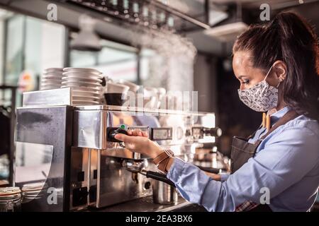 La maschera che indossa la cameriera aziona la macchina del caffè in un caffè. Foto Stock