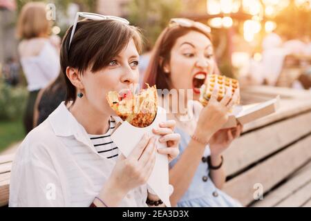 Due felici amici donne che si rilassano nel parco cittadino e mangiano cucina moderna di strada. Dieta sana e vacanza fastfood urbano Foto Stock