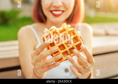 La donna felice in un parco cittadino sta pranzando e tenendo un sandwich viennese di cialde. Cibo di strada moderno, delizioso e ad alta caloria Foto Stock