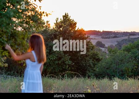 Profilo di tramonto offuscato di una ragazza con bionda lunga capelli vestiti in bianco mentre tocca le foglie di l'albero magico Foto Stock