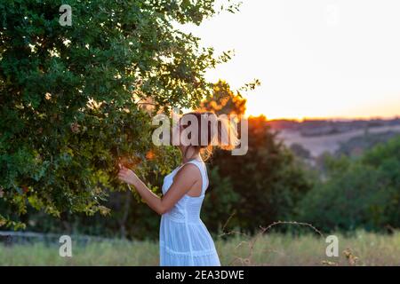 Profilo di una giovane ragazza in abito bianco al tramonto con capelli biondi legati vicino all'albero magico Foto Stock