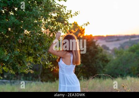 Profilo di una giovane ragazza con lunghi capelli biondi vestiti in bianco con la testa girata verso il tramonto come tocca le foglie dell'albero magico Foto Stock
