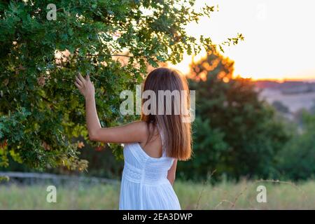 Profilo di una giovane ragazza con lunghi capelli biondi vestiti in bianco con la testa girata verso il tramonto come tocca le foglie dell'albero magico Foto Stock