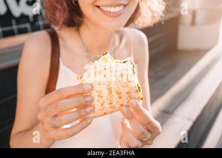 La donna felice mangia un pane piatto fast food con ripieno seduto in strada e riposandosi dopo una giornata di lavoro. Concetto di cibo sano e calorie extra Foto Stock