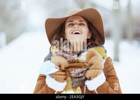 sorridente donna moderna di 40 anni in cappello marrone e sciarpa con mittens e maschera medica in camice di pelle di pecora all'aperto nel parco della città in inverno. Foto Stock