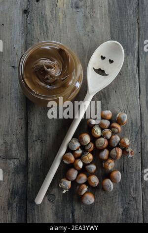 Vaso con pasta di nocciole, cucchiaio di legno con viso e nocciole secche su fondo di legno Foto Stock