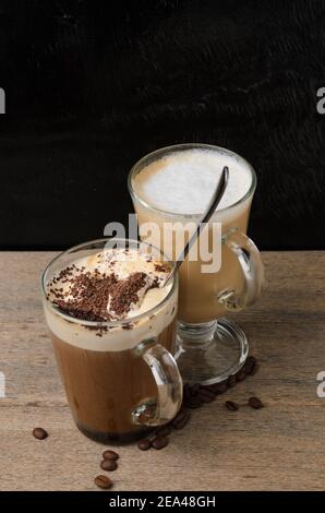 Due tazze in vetro con cappuccino e cioccolato ghiacciato su un tavolo di legno con fondo a trama nera. Messa a fuoco selettiva. Foto Stock