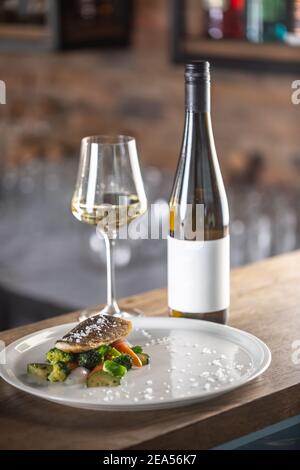 Pesce sano cucinato con verdure al vapore servito su un piatto accanto ad una bottiglia di vino bianco. Foto Stock