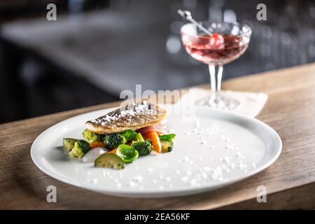 Pesce sano cucinato con verdure al vapore servito su un piatto accanto ad un cocktail negroni. Foto Stock