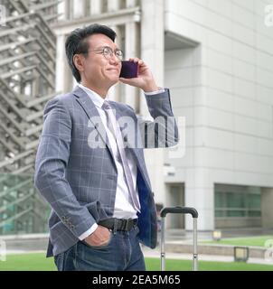 Uomo d'affari asiatico con cravatta e giacca, sorridente e parlando al telefono con edifici moderni sullo sfondo. Foto Stock