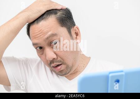 Un uomo di mezza età splende nello specchio e si preoccupa di perdita di capelli o grigio dei capelli isolato uno sfondo bianco, concetto di assistenza sanitaria Foto Stock