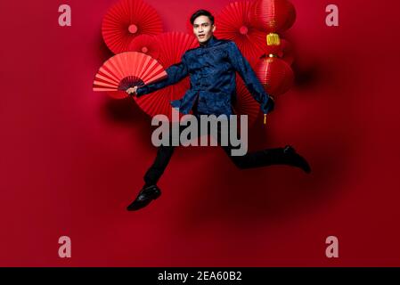 Uomo asiatico in costume tradizionale che tiene fan e salta dentro Stile orientale decorato sfondo rosso per i concetti cinesi di nuovo anno Foto Stock