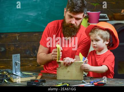 Padre, genitore con barba che insegna a piccolo figlio a segare mentre il figlio gioca con sega giocattolo. Ragazzo, bambino occupato in casco protettivo imparare ad usare la sega a mano con papà. Concetto di giochi educativi. Foto Stock