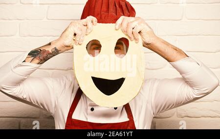 Concetto di processo di cottura. Chef prepara la pizza. Cuocete con il viso nascosto in borgogna uniforme giochi con l'impasto. L'uomo con la barba tiene l'impasto arrotolato con il viso smiley tagliato su sfondo bianco della parete in mattoni. Foto Stock