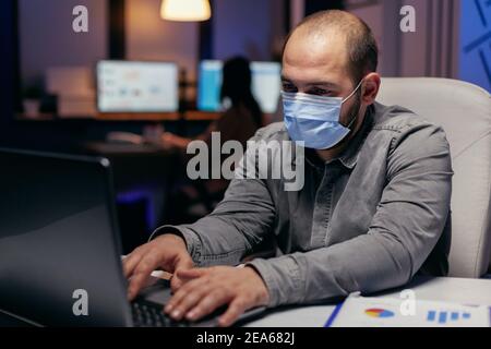 Il giovane uomo d'affari indossa la maschera sul posto di lavoro per prevenire le infezioni. L'uomo stressato in corporazione che lavora duro per rifinire un progetto che indossa la maschera di faccia come precauzione di sicurezza dovuto la pandemia del coronavirus. Foto Stock