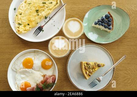 Colazione luminosa su sfondo di legno marrone, vista dall'alto. Dessert gustosi, omelette, uova fritte e tazza di caffè. Foto Stock