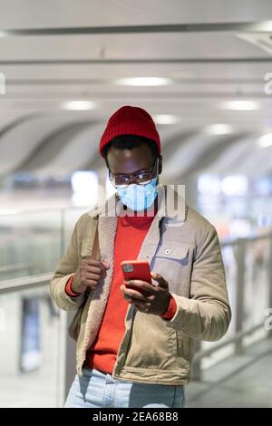 Uomo nero che usa lo smartphone nel terminal dell'aeroporto, indossare la maschera medica facciale, utilizzando il cellulare. Covid-19. Foto Stock