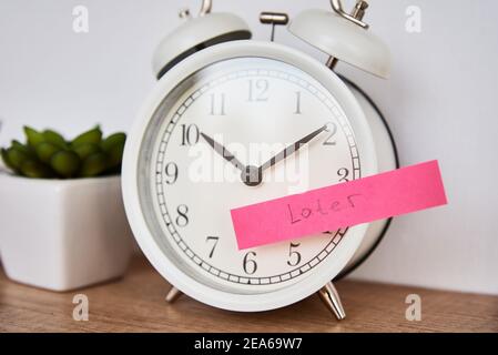 Concetto di procrastinazione e ritardo. Nota appiccicosa con parola più tardi sulla sveglia bianca. Tempo di urgenza Foto Stock
