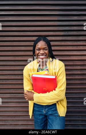 Foto di scorta di giovane ragazza nera che tiene il notebook, sorridendo e guardando la fotocamera. Foto Stock