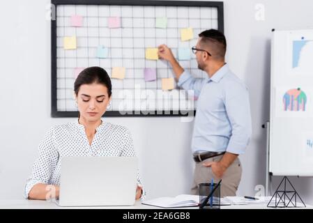 donna d'affari che usa il laptop vicino a un collega che mette a bordo note appiccicose e sfondo sfocato Foto Stock