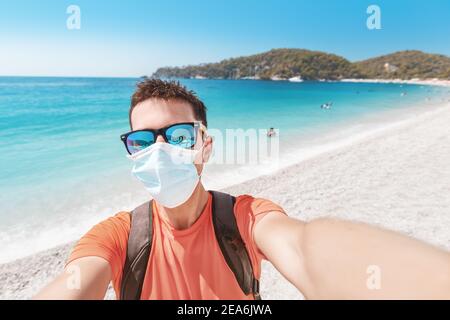 Un turista maschile che indossa occhiali da sole e una maschera medica protettiva Passeggiate e poggia su una spiaggia di sabbia sul Mediterraneo mare durante un pandemi coronavirus Foto Stock