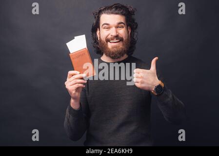 Affascinante giovane uomo con la bearded in casual che tiene i biglietti del passaporto e mostra il pollice in su. Foto Stock