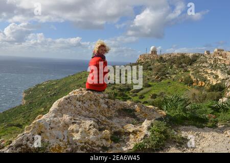 Vista posteriore di una donna seduta su una roccia che guarda le scogliere di Dingli con la stazione radar di Dingli in lontananza, Malta Foto Stock