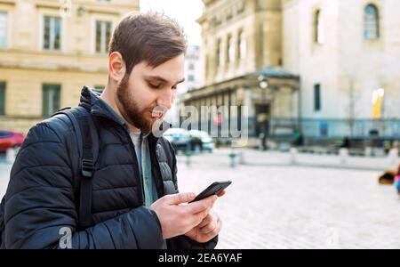 Giovane elegante e accattivante che guarda sullo smartphone. Uomo in strada all'aperto in Europa utilizzando il telefono cellulare. Foto Stock