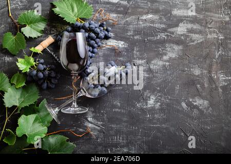 Bicchieri da vino pieni di vino rosso e uva. Bottiglie di vino, grappoli d'uva con foglie e tappi di vite su fondo rustico scuro di cemento. Vino piatto Foto Stock