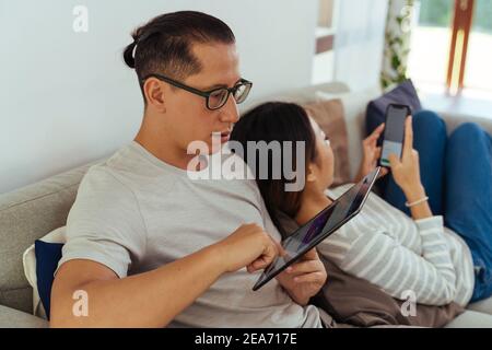 Anni 30 giovane adulto asiatico uomo e donna giocare un telefono cellulare insieme si siede sul divano in un accogliente soggiorno a casa. Felice utente di Internet di coppia in abiti casual sul divano. Concetto di utilizzo della tecnologia in Asia Foto Stock