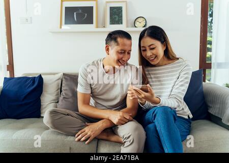 Anni 30 giovane adulto asiatico uomo e donna giocare un telefono cellulare insieme si siede sul divano in un accogliente soggiorno a casa. Felice utente di Internet di coppia in abiti casual sul divano. Concetto di utilizzo della tecnologia in Asia Foto Stock