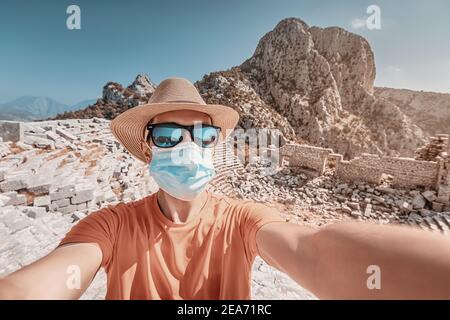 Un viaggiatore maschile indossa una maschera medica e occhiali da sole con un cappello e prende un selfie contro le rovine dell'antico anfiteatro di Termessos. Quaranti Foto Stock