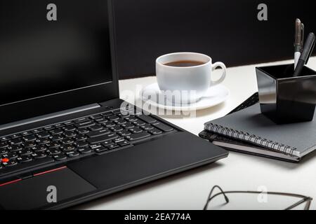 Computer portatile nero su sfondo bianco. Bicchieri, tazza di caffè, forniture per ufficio. Spazio di lavoro domestico per lavoro o studio all'interno dell'ufficio Foto Stock