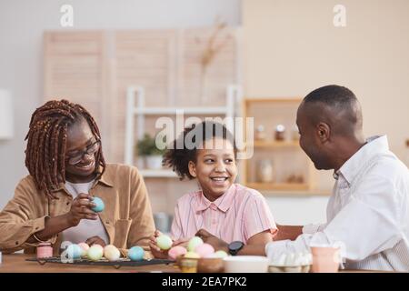 Ritratto di ridere famiglia afroamericana dipingendo insieme le uova di Pasqua seduto al tavolo di legno in casa accogliente interno e godendo Arte fai da te Foto Stock