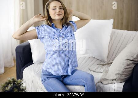 Donna bionda felice messo le mani dietro la testa seduta appoggiata sul divano femmina caucasica godere pigro week-end o vacanza in accogliente soggiorno, casalinga Foto Stock