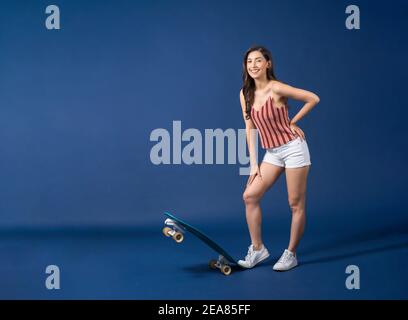 Felice giovane donna asiatica in piedi sul surfskate o skateboard su sfondo blu colore, esercizio e sport, trendy e extream concetto sportivo Foto Stock