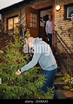 Uomo di mezza età che mette le luci della fata sull'albero di Natale all'aperto per la donna anziana, Svezia. Concetto di stile di vita, cura, celebrazione Foto Stock