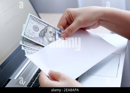 Busta con dollari USA in mani femminili. Donna tira i soldi da una busta sullo sfondo del laptop, salari, bonus o concetto di tangente Foto Stock