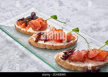 tre bruschette con trota, salmone, formaggio cremoso e microsverde su un piatto di vetro su fondo bianco Foto Stock