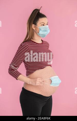 Giovane donna incinta che indossa una maschera facciale che tiene un'altra maschera sopra il ventre esposto su uno sfondo rosa.concetto di gravidanza e sicurezza. Foto Stock