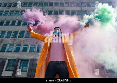 Il giovane uomo in maschera respiratore regge bombe di fumo in mani alzate. Protesta, libertà, concetto di rivoluzione Foto Stock
