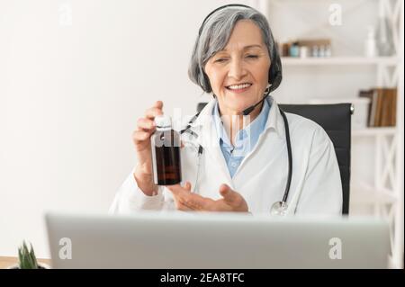 Una vecchia dottoressa anziana o un farmacista con un auricolare in un camice da laboratorio seduto di fronte a un computer portatile, mostrando un flacone di medicinale prescritto da Foto Stock