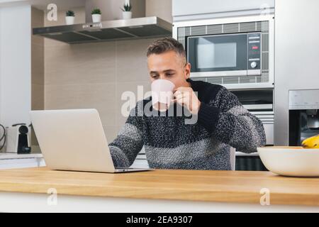 Giovane uomo che usa un notebook mentre fa colazione, legge, scrive e-mail o lavora online seduto in cucina moderna Foto Stock