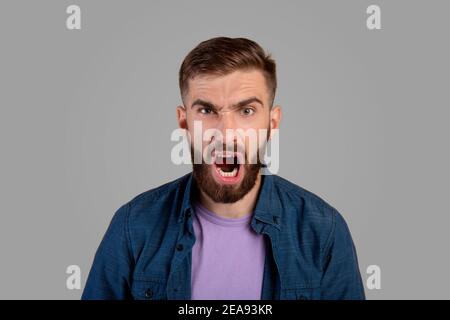 Ritratto di giovane ragazzo bearded gridando furiosamente, esprimendo la sua rabbia su sfondo grigio studio. Emozioni negative Foto Stock