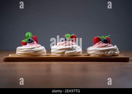 Tre mini torte Pavlova con ripieno di crema di fragole decorate con frutti di bosco freschi e foglie di menta su superficie di legno. Isolato. Sfondo grigio Foto Stock