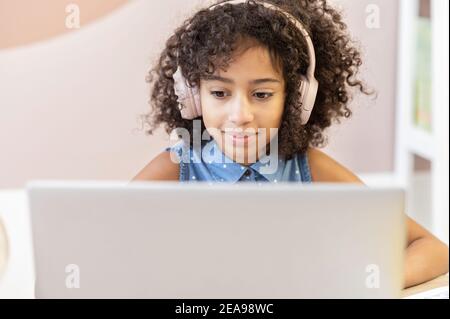 Una studentessa africana piuttosto curly che indossa una cuffia usando un laptop per studiare a distanza, una ragazza biracial graziosa guarda lo schermo, facendo Foto Stock