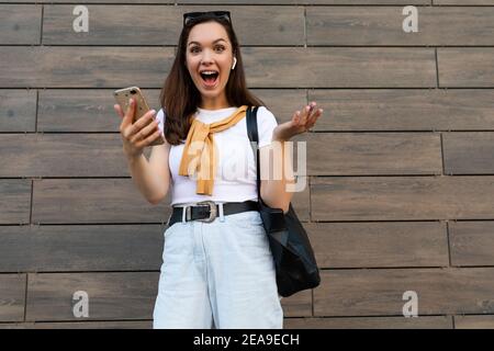 Foto di attraente scioccata sorpresa giovane donna che indossa abiti casual in piedi per strada utilizzando il telefono cellulare guardando fotocamera Foto Stock