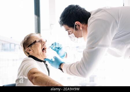 Vista laterale del medico maschile in maschera protettiva che assume COVID 19 test da parte di una paziente anziana di sesso femminile a casa Foto Stock