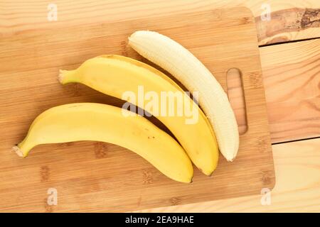 Due banane biologiche gialle mature succose e una sbucciata su un tagliere di bambù, primo piano, su uno sfondo di legno naturale. Foto Stock