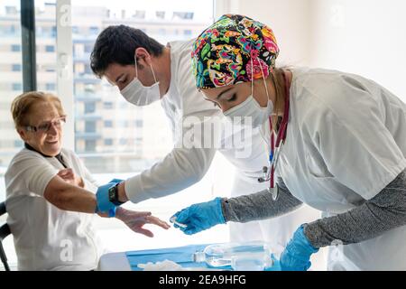 Vista laterale dei medici in maschere protettive che prelevano campioni di sangue da anziana donna seduta in camera a casa Foto Stock